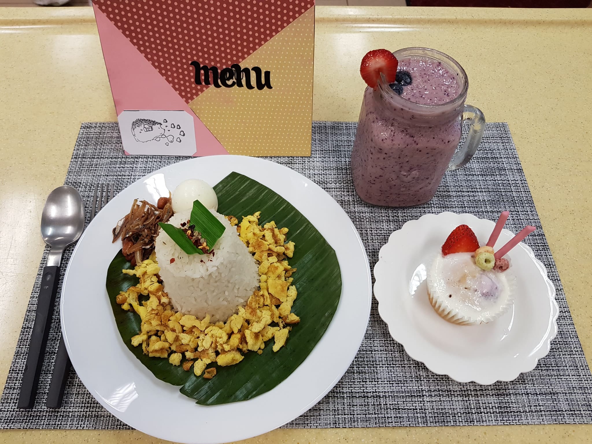 A meal set with rice and scrambled eggs on a banana leaf, a berry smoothie, and a decorated cupcake on a tray.